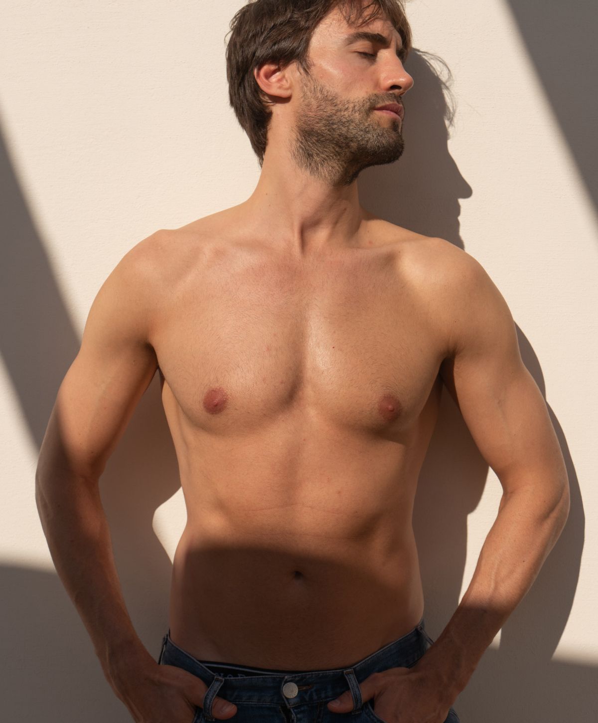 Shirtless man leaning against wall with shadows.